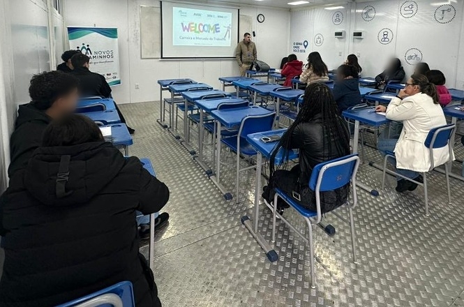 Foto horizontal: jovens participam de atividade formativa em sala de aula do programa Novos Caminhos, acompanhando apresentação projetada ao fundo.