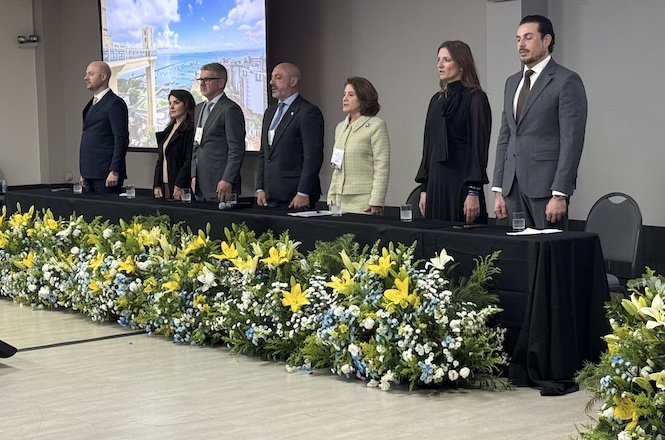 Foto horizontal: autoridades compõem mesa de honra durante solenidade institucional, diante de arranjo floral e painel de apresentação ao fundo.