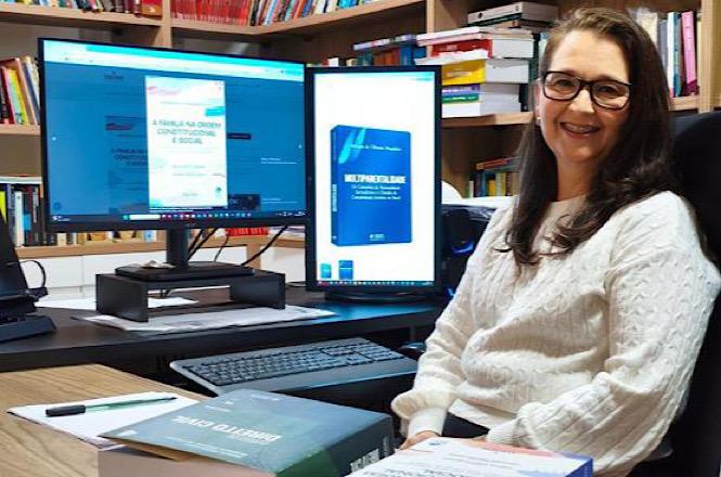Foto horizontal: autora posa em escritório cercada por livros e telas com sua obra em destaque, durante divulgação de publicação jurídica.