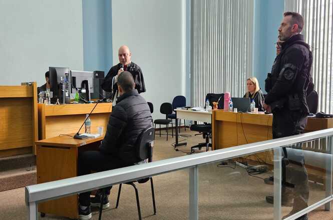 Foto horizontal: audiência judicial ocorre em sala de tribunal, com réu sentado diante do magistrado e agentes de segurança acompanhando o procedimento.