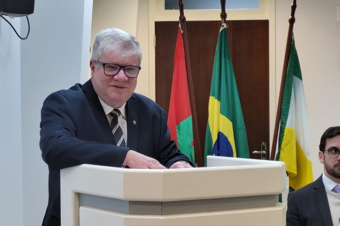 Foto horizontal: juiz aposentado Renato Bratti discursa em solenidade institucional, diante das bandeiras de Santa Catarina e do Brasil.