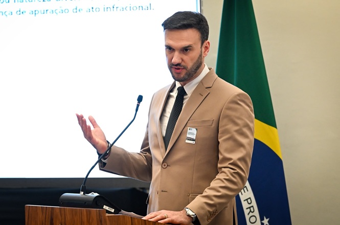 Foto horizontal: magistrado profere palestra em evento institucional, ao lado da bandeira do Brasil, durante apresentação sobre temas ligados à infância e juventude.