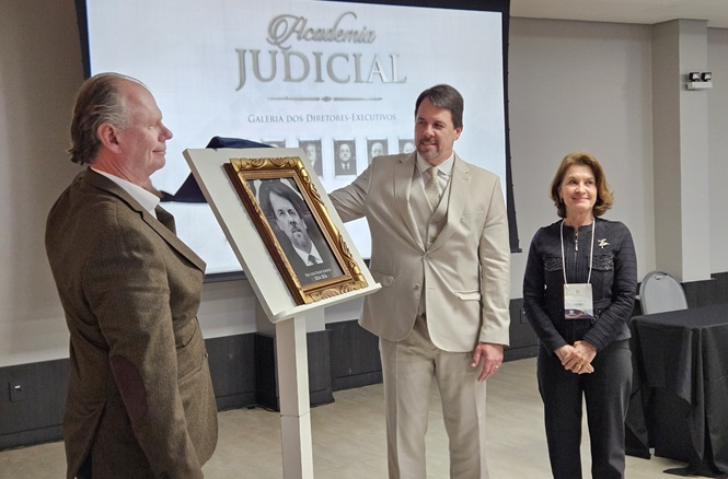 Ambiente interno com tela ao fundo exibindo “Academia Judicial” e “Galeria dos Diretores Executivos”. Em primeiro plano, três pessoas estão em pé, ao lado de um pedestal branco com um quadro emoldurado: à esquerda, Rubens Schulz, de perfil voltado ao centro; no meio, Luiz Felipe Schuch apresenta o quadro com foto em preto e branco; à direita, Vera Copetti está próxima a uma mesa com toalha escura.