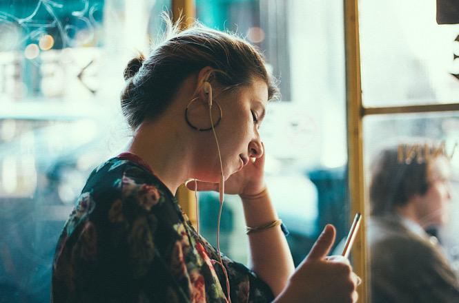 Foto ilustrativa: mulher utiliza smartphone com fones de ouvido em ambiente público, simbolizando uso de tecnologia, conectividade digital e consumo de conteúdo online.