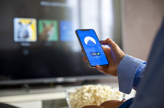 Foto ilustrativa: pessoa utiliza aplicativo de VPN em smartphone diante de televisão conectada, simbolizando privacidade digital, segurança da informação e proteção de dados na internet.
