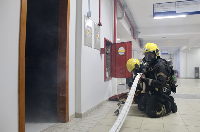 Foto horizontal: bombeiros militares equipados para combate a incêndio atuam em simulado de emergência dentro de prédio institucional, utilizando mangueira e equipamentos de proteção respiratória.