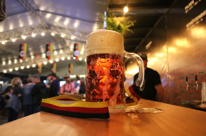 Foto ilustrativa: caneca de cerveja sobre mesa em ambiente festivo decorado com cores da Alemanha, remetendo a celebrações culturais, gastronomia e tradições germânicas.