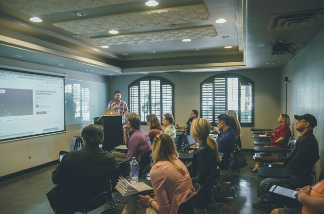 Foto horizontal: participantes acompanham apresentação em sala de treinamento durante curso de capacitação, com exposição de conteúdo projetado e interação entre instrutor e público.