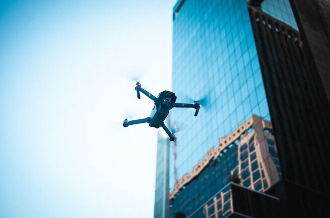 Foto ilustrativa: drone sobrevoa área urbana entre edifícios, representando o uso de aeronaves remotamente pilotadas em atividades de monitoramento, inspeção, segurança e produção de imagens.