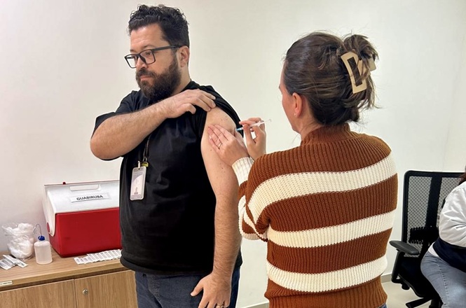 Foto horizontal: servidor recebe vacina aplicada por profissional da saúde durante campanha de imunização no ambiente de trabalho.
