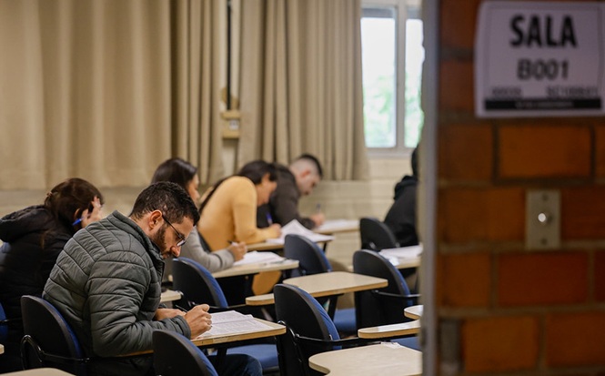 Foto horizontal: participantes realizam prova em sala de aula durante processo seletivo ou atividade de capacitação, concentrados no preenchimento das avaliações.