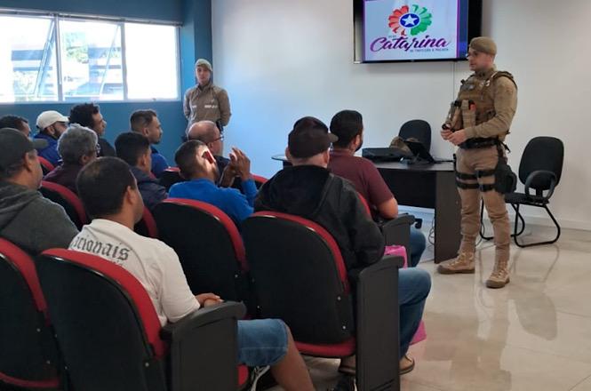 Foto horizontal: participantes acompanham palestra educativa em auditório, enquanto palestrante conduz atividade de orientação e prevenção, com apoio de equipe técnica e recursos audiovisuais.
