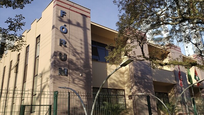 Foto horizontal: prédio bege com letras pretas “FÓRUM”, árvores verdes e céu azul, urbano, destacando fachada institucional