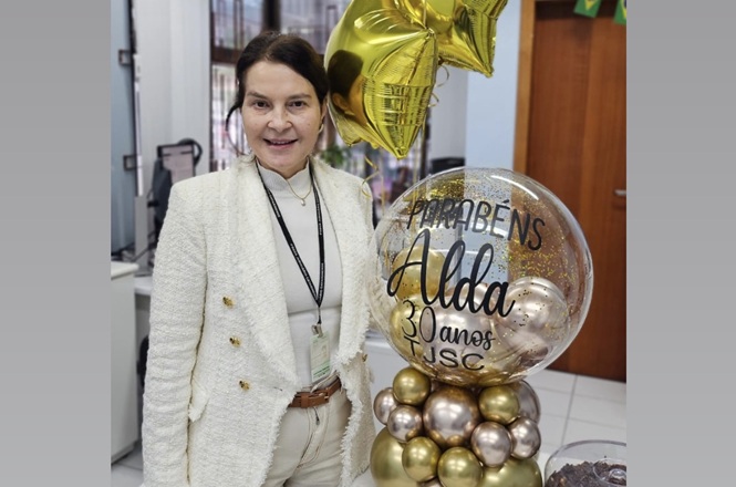 Foto vertical: servidora posa ao lado de balões comemorativos em homenagem aos seus 30 anos de atuação no TJSC.