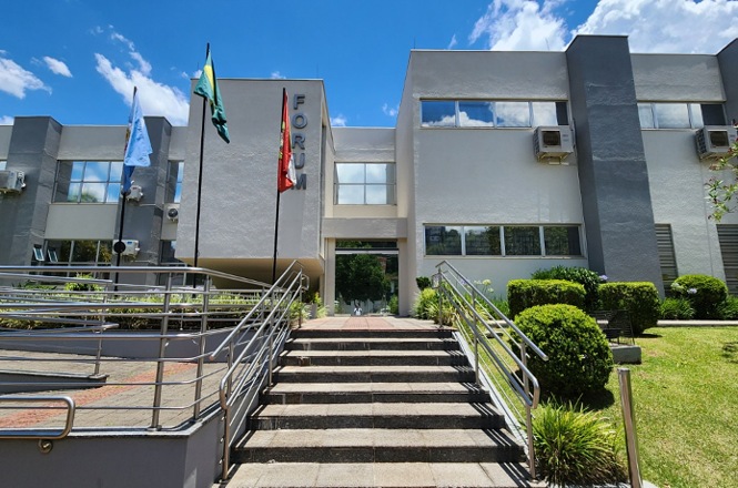Foto horizontal: fachada do Fórum da comarca, com escadaria de acesso, rampa de acessibilidade e mastros com bandeiras.
