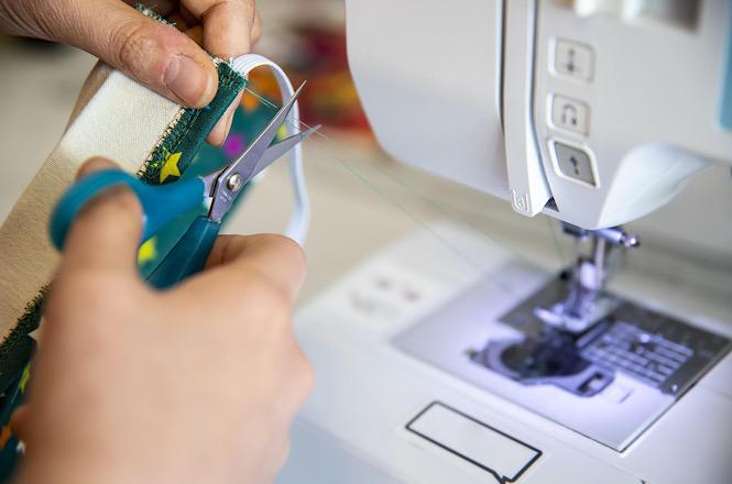 Foto horizontal: mãos cortam tecido ao lado de máquina de costura, representando atividade de confecção e trabalho artesanal.