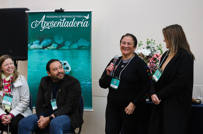 Foto horizontal: participante fala ao microfone durante atividade do Programa de Preparação para a Aposentadoria, acompanhada por facilitadores.