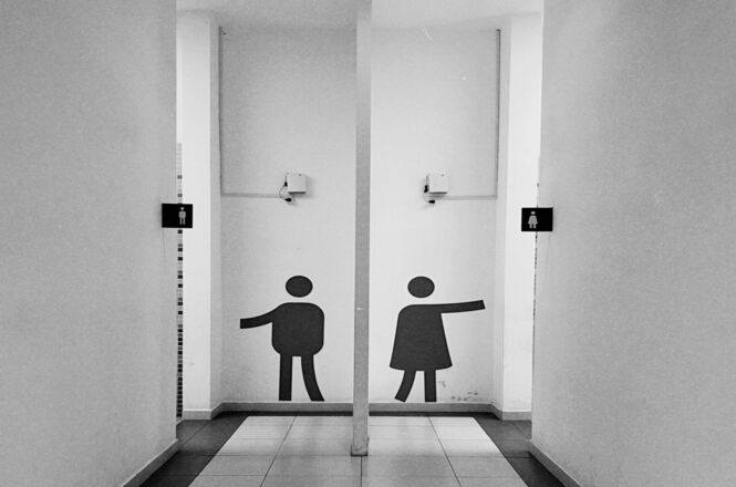 Foto corredores brancos com símbolos pretos de banheiro, ambiente interno, sinalizando sanitários masculino e feminino