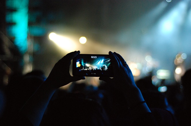 Foto colorida: pessoa gravando show com celular, auditório escuro iluminado, evento musical, registrando apresentação em azul, preto e branco