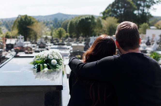 Foto colorida: duas pessoas diante de túmulo florido, cemitério ao ar livre, contexto de luto, prestando homenagem em verde, branco e preto