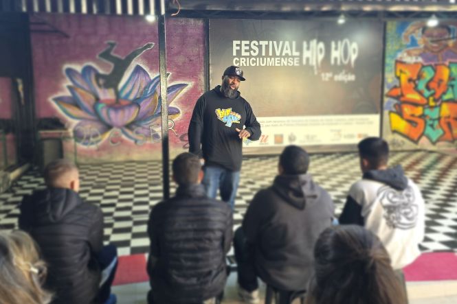 Foto horizontal: homem conduz roda de conversa durante atividade do Festival Criciumense de Hip Hop, com participantes sentados à sua frente e grafites ao fundo.