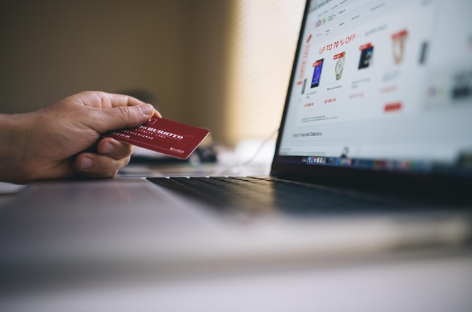 Foto horizontal: mão segura cartão bancário diante de notebook com página de compras on-line, representando transações e comércio eletrônico.