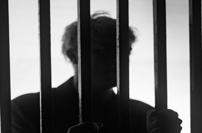 Foto horizontal: silhueta de pessoa atrás de grades, segurando as barras com as mãos, em imagem que simboliza privação de liberdade e sistema prisional.