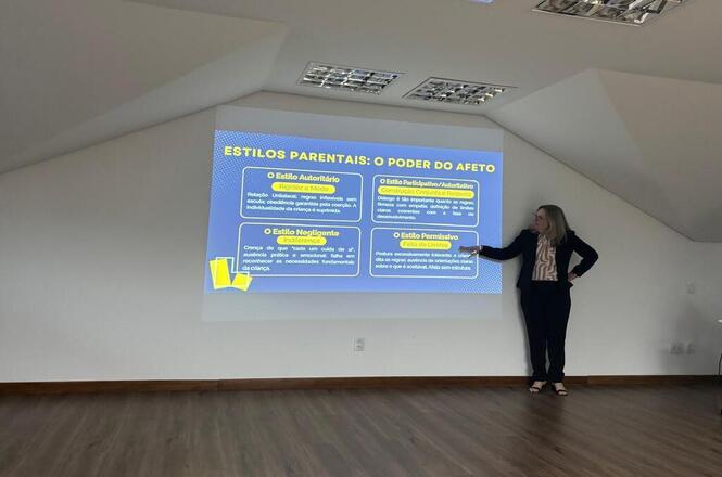 Foto horizontal: palestrante apresenta conteúdo sobre estilos parentais durante capacitação, com projeção de slides em sala de treinamento.