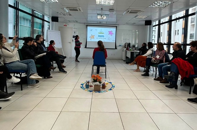 Foto horizontal: participantes acompanham atividade em sala de capacitação, com apresentação projetada e dinâmica em grupo organizada em círculo.