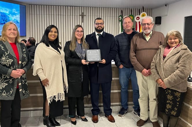 Foto horizontal: grupo posa durante homenagem a representante da comunidade, com entrega de placa de reconhecimento em sessão solene.