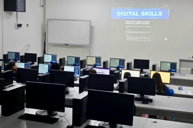 Foto horizontal: participantes acompanham aula em laboratório de informática, com computadores individuais e conteúdo projetado sobre competências digitais.