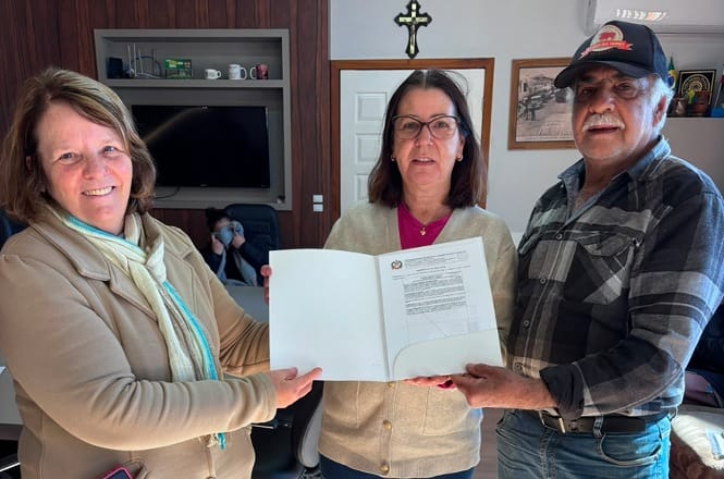 Foto horizontal: três pessoas exibem documento oficial após assinatura, durante ato de formalização realizado em ambiente institucional.