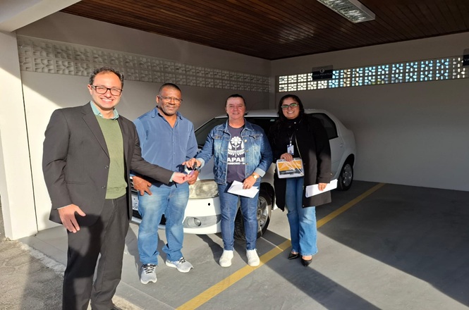 Foto horizontal: quatro pessoas posam ao lado de veículo durante cerimônia de entrega de automóvel, com um participante recebendo a chave e documentos.