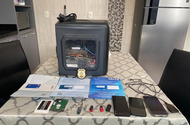 Foto horizontal: computador, celulares, dispositivos de armazenamento, documentos e outros objetos apreendidos estão organizados sobre uma mesa durante ação policial.