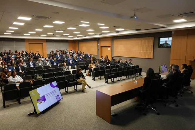 Horizontal: Auditório com público e mesa de palestrantes, em ambiente corporativo com iluminação interna ampla.