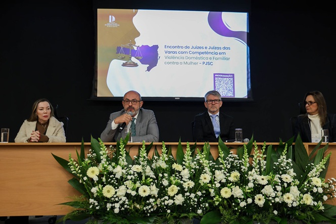 Mesa de autoridades em um auditório, com quatro pessoas. À frente da mesa, há um grande arranjo de flores brancas. Ao fundo, um telão exibe a apresentação do evento “Encontro de Juízes e Juízas das Varas com Competência em Violência Doméstica e Familiar contra a Mulher – PJSC”.