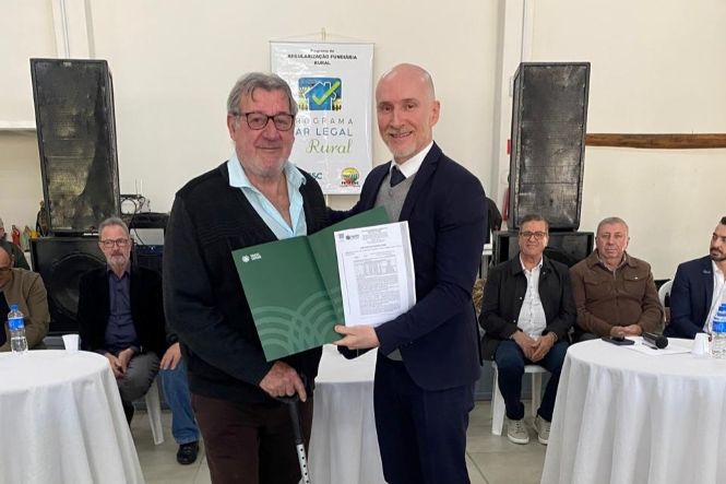 Foto horizontal: participante recebe documento de regularização fundiária durante cerimônia do Programa Lar Legal Rural, diante de autoridades e convidados.