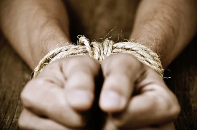 Foto horizontal: mãos de pessoa amarradas por corda, em imagem ilustrativa que representa cárcere privado, sequestro, violência e privação de liberdade.