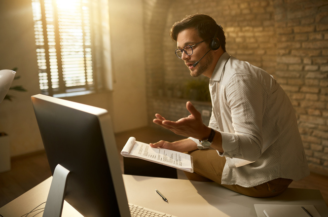 Foto horizontal: homem utiliza computador e headset durante videoconferência ou atendimento remoto, consultando anotações enquanto conversa em ambiente de trabalho.