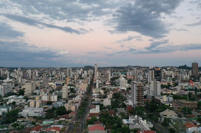 Foto horizontal: vista aérea de área urbana com edifícios residenciais e comerciais, avenidas e bairros distribuídos sob céu parcialmente nublado, em imagem panorâmica da cidade.