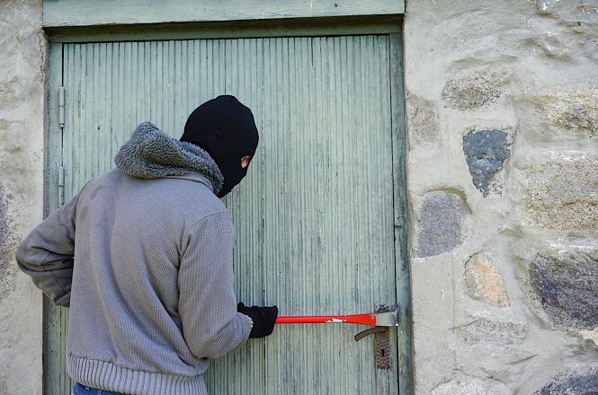 Foto horizontal: pessoa com o rosto coberto utiliza pé de cabra para arrombar a porta de um imóvel, em imagem ilustrativa que representa furto, invasão de domicílio e crimes contra o patrimônio.