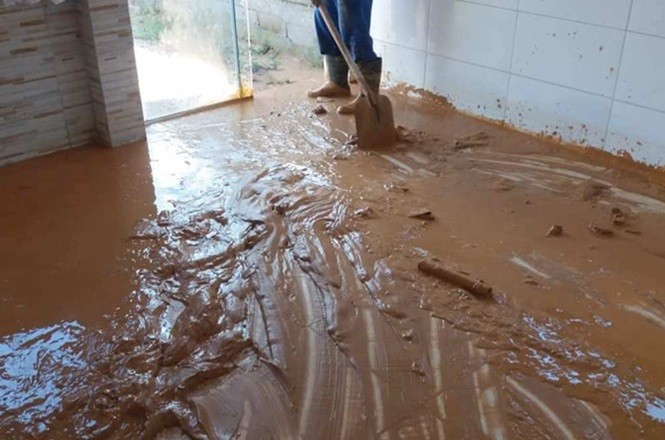 Foto horizontal: pessoa utiliza pá para remover lama acumulada no interior de imóvel após alagamento, em cena que retrata os impactos de enchentes e o processo de limpeza.
