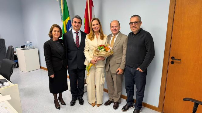 Fotografia em ambiente interno, com parede cinza ao fundo e as bandeiras do Brasil e de Santa Catarina parcialmente visíveis. Da esquerda para a direita, estão a desembargadora Vera Copetti, vestindo tailleur preto; o desembargador Leoni Martins, de terno escuro, camisa branca e gravata em tons de vinho; ao centro, a magistrada Alexandra Lorenzi, sorrindo e segurando um buquê de flores, vestida com conjunto em tom claro; ao seu lado, o professor Marcelo Dantas, de terno bege com gravata dourada; e, por fim, Leandro Aguiar, usando suéter escuro, calça jeans e óculos. Todos estão lado a lado, voltados para a câmera, em um momento de celebração após a defesa de doutorado. À direita da imagem, há uma porta de madeira.