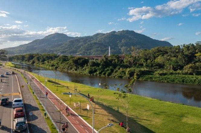Vista aérea mostra rio, ciclovia, avenida e área verde aos pés de morros sob céu com poucas nuvens.