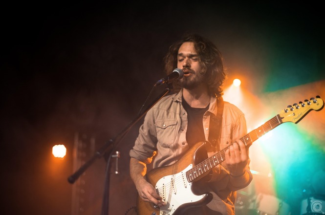 Cantor se apresenta com guitarra e microfone em show, sob iluminação cênica e efeitos de fumaça.