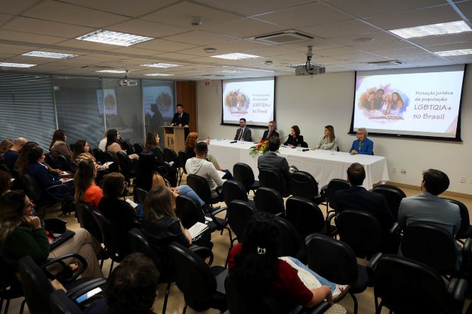 Fotografia em plano aberto de um auditório durante o seminário "Proteção Jurídica da População LGBTQIA+ no Brasil". Em primeiro plano, participantes estão sentados em cadeiras pretas, voltados para a mesa de autoridades posicionada à frente da sala. Ao fundo, um palestrante faz uso da tribuna enquanto, à direita, seis integrantes compõem a mesa principal, coberta por toalha branca e decorada com um arranjo de flores ao centro. Dois telões projetam a identidade visual do evento, com a imagem de um grupo de pessoas e o título do seminário. O ambiente é amplo, iluminado por luzes no teto, com paredes claras e janelas na lateral esquerda.