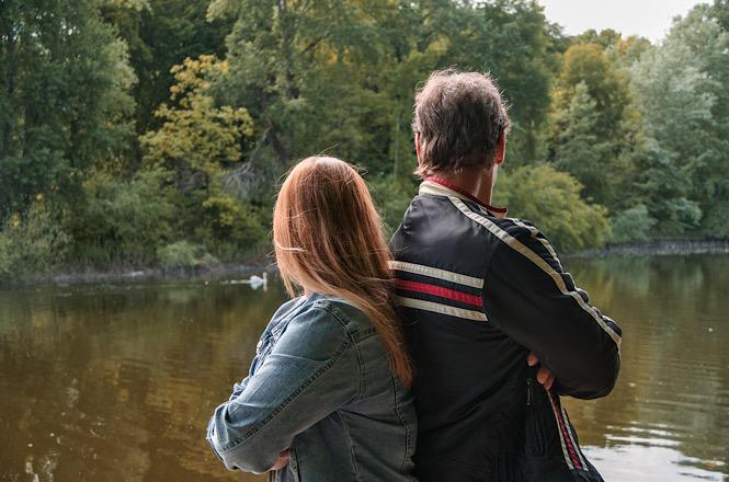 Casal observa lago cercado por árvores, de costas para a câmera, em momento de contemplação da natureza.