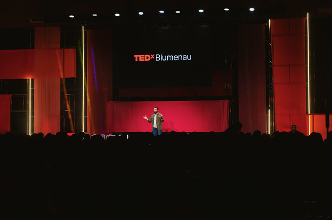 Palestrante se apresenta no palco durante o TEDxBlumenau diante de plateia em auditório lotado.