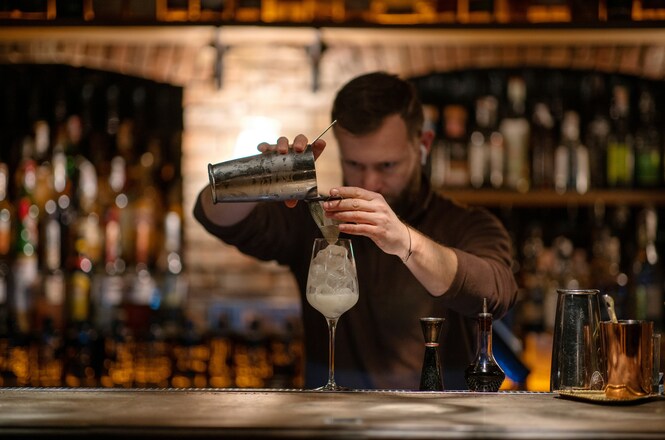 Bartender prepara drinque em balcão de bar, utilizando coqueteleira e taça com gelo.