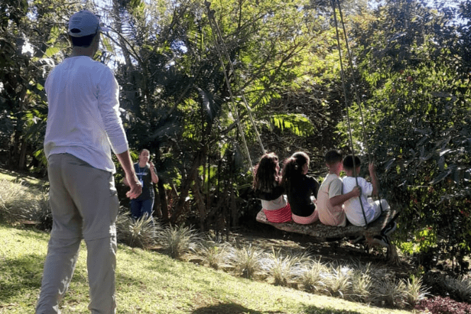 Crianças brincam em balanço enquanto adulto acompanha a atividade em área verde arborizada.